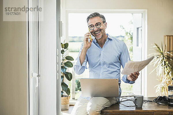 Glücklicher Geschäftsmann mit Dokument  der im Heimbüro auf dem Smartphone spricht