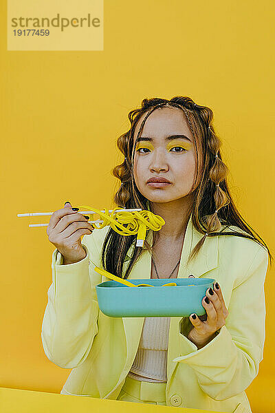 Junge Frau isst verkabelte Nudeln aus einer Bento-Box vor gelbem Hintergrund