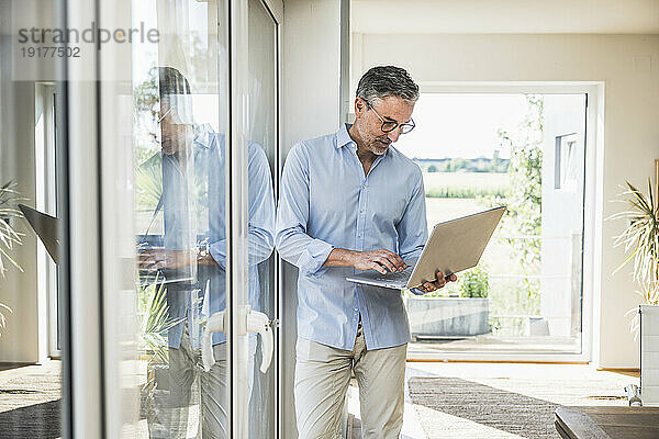 Geschäftsmann arbeitet am Laptop und lehnt am Fenster im Heimbüro