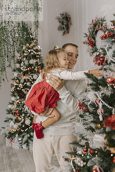 Lächelnder Vater trägt Tochter und berührt Weihnachtsbaum