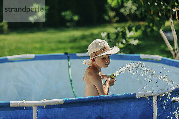 Junge mit Hut spielt an sonnigem Tag mit Wasserpfeife im Planschbecken