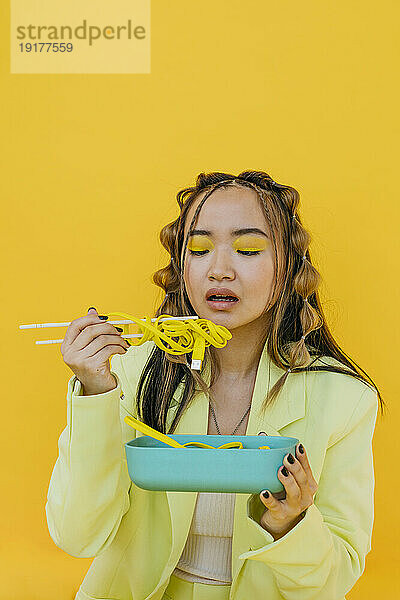 Young woman eating noodles like wire from bento box against yellow background