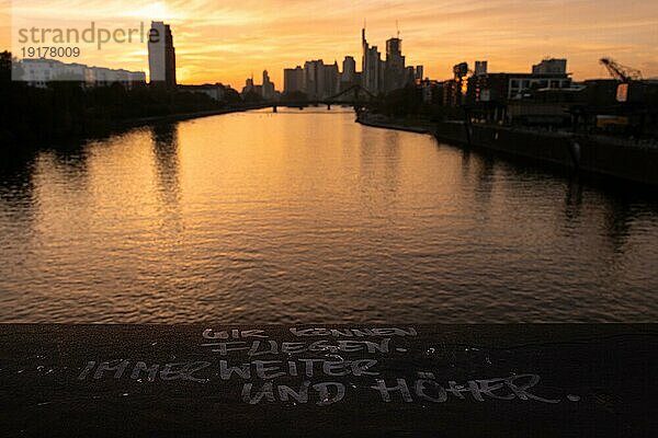 WIR KÖNNEN FLIEGEN. IMMER WEITER UND HÖHER. steht auf einem Geländer der Deutschherrnbrücke im Frankfurter Osthafengebiet  während sich im Hintergrund nach Sonnenuntergang die Silhouette der Frankfurter Bankenskyline abzeichnet.  Osthafen  Frankfurt am Main  Hessen  Deutschland  Europa