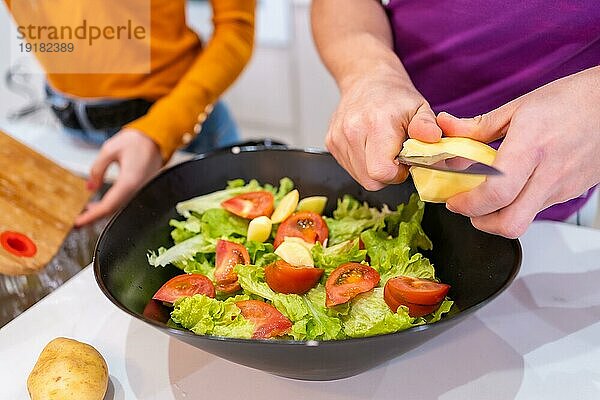 Horizontales Foto mit Nahaufnahme eines Mannes  der neben seiner Partnerin zu Hause Zutaten für einen Salat schneidet