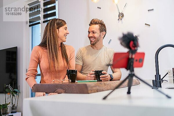 Frontalaufnahme eines glücklichen jungen Paares  das von zu Hause aus online streamt und dabei Kaffee trinkt