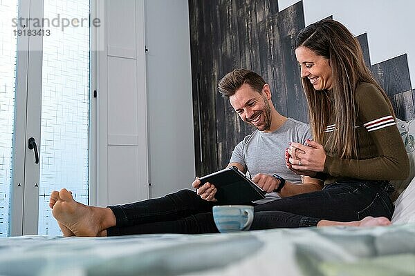 Seitenansicht eines glücklichen Paares  das mit einem digitalen Tablet und einer Kaffeetasse Zeit auf dem Bett verbringt