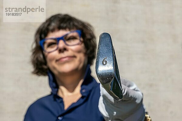 Glückliche Golferin mit Brille hält ihren 6er Golfschläger hoch und schaut in die Kamera an einem sonnigen Tag in der Schweiz