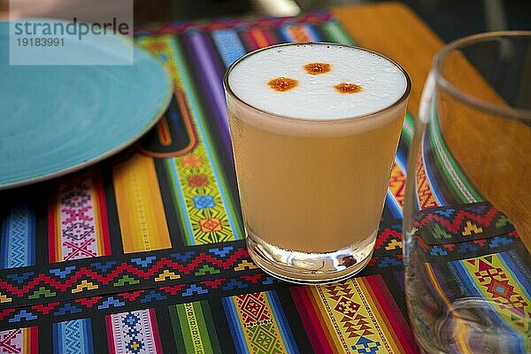 Pisco Sour alkoholisches Getränk  peruanischer Cocktail  hergestellt aus Pisco (peruanischer Traubenschnaps)  Limettensaft  Sirup und Eiweiß  im Glas auf Tischsets mit traditionellen Mustern. Selektiver Fokus  unscharfer Hintergrund  Fokus auf das vordere Glas und den Schaum
