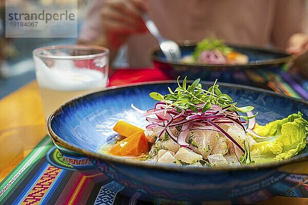 Frische Ceviche  serviert in einem blauen Teller auf einem Tischset mit traditionellen Mustern  Pisco Sour-Cocktail. Unerkennbarer Erwachsener im unscharfen Hintergrund. Ceviche nach peruanischer Art aus rohem Fisch  roten Zwiebeln  Mais  geröstetem Mais und Süßkartoffeln. Selektiver Fokus