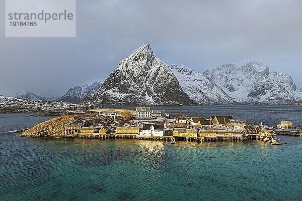 Traditionelle gelbe Rorbuer Hütten auf der Insel Sakrisøy  hinten schneebedeckte felsige Berge  Sakrisoy  Reine  Lofoten  Norwegen  Europa