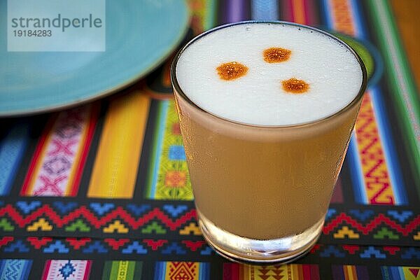 Pisco Sour alkoholisches Getränk  peruanischer Cocktail  hergestellt aus Pisco (peruanischer Traubenschnaps)  Limettensaft  Sirup und Eiweiß  im Glas auf Tischsets mit traditionellen Mustern. Selektiver Fokus  unscharfer Hintergrund  Fokus auf das vordere Glas und den Schaum