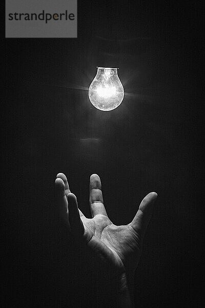Hand greift von unten nach leuchtender Glühbirne  Schwarzweiß  künstlerisch