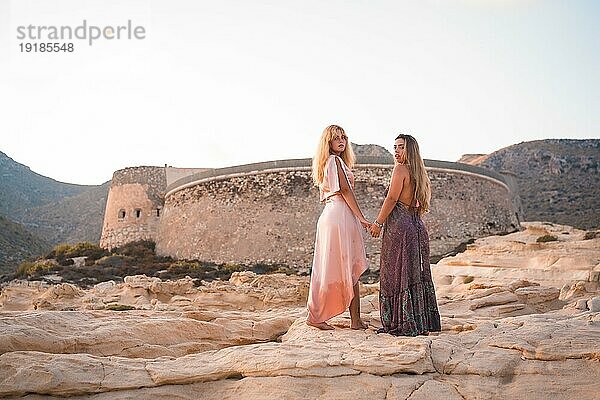 Ein paar kaukasische Mädchen mit rosa Kleidern  im Sommer am Playazo de Rodalquilar in Cabo de Gata neben der Burg im Sommer