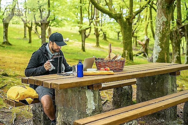 Ein junger kaukasischer Vater bei der Telearbeit an einem Picknicktisch mit Computer im Wald. Sommerlicher Lebensstil