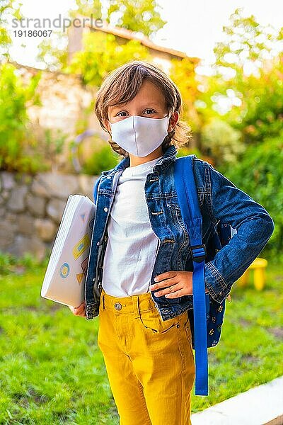 Kaukasisches Kind mit Gesichtsmaske  bereit für die Rückkehr zur Schule. Neue Normalität  soziale Distanz  Coronaviruspandemie Covid 19. Mit Jeansjacke  Rucksack und Notizblock in der Hand
