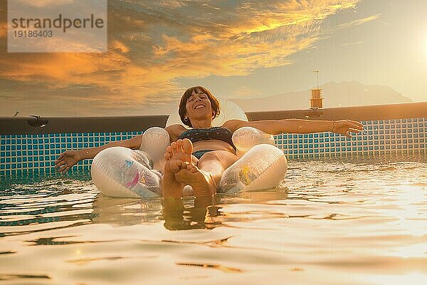 Eine junge Brünette Kaukasier lächelnd genießen den Sommer auf einer Matte im Pool  bei Sonnenuntergang in der goldenen Stunde