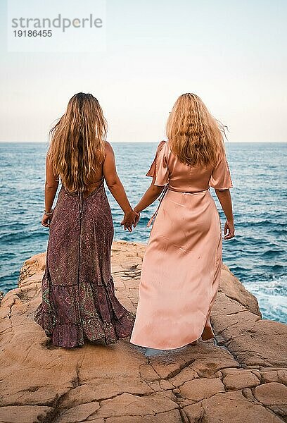 Ein paar kaukasische Mädchen in schönen Kleidern  die sich am Meer im Sommer liebevoll an den Händen halten