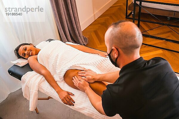 Massage zu Hause  Durchführung einer Massage zu Hause zufriedenen Kunden auf die Beine. Mit einer Gesichtsmaske in der Coronaviruspandemie