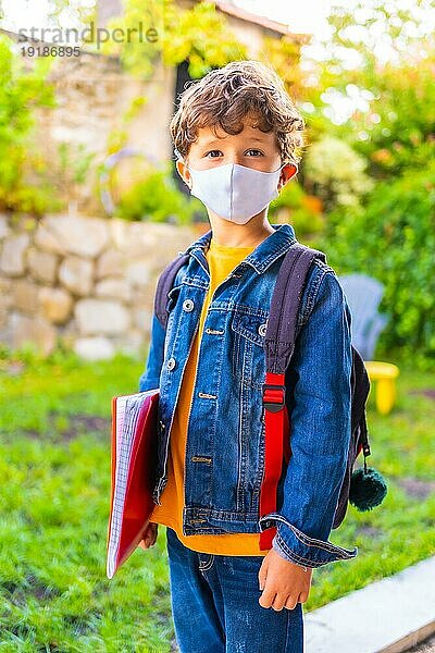 Kaukasisches Kind mit Gesichtsmaske  bereit für die Rückkehr zur Schule. Neue Normalität  soziale Distanz  Coronaviruspandemie Covid 19. Jacke  Rucksack und ein roter Block für Notizen in der Hand
