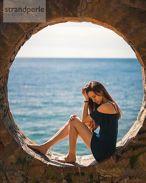 Ein junges hübsches rothaariges kaukasisches Mädchen in einem blauen Kleid lächelnd am Meer sitzend