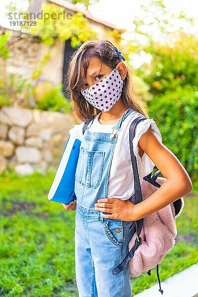 Kaukasisches Mädchen mit Gesichtsmaske  bereit  wieder zur Schule zu gehen. Neue Normalität  soziale Distanz  Coronaviruspandemie Covid 19. Jacke  Rucksack und ein blauer Block für Notizen in der Hand