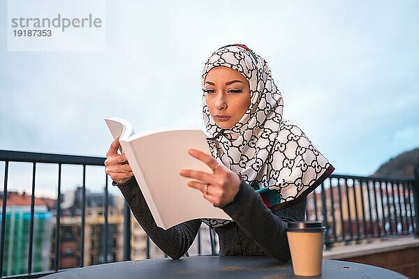 Arabisches Mädchen mit weißem Schleier am Computer auf der Terrasse eines Coffeeshops  ein schönes Buch lesend