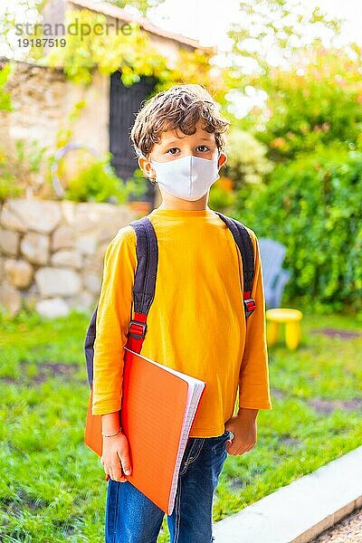 Kaukasisches Kind mit Gesichtsmaske  bereit für die Rückkehr zur Schule. Neue Normalität  soziale Distanz  Coronaviruspandemie Covid 19. Orangefarbenes TShirt Rucksack und ein Notizblock in der Hand