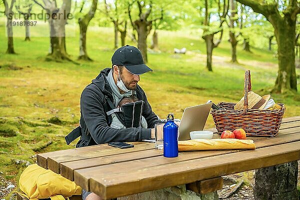 Ein junger kaukasischer Vater bei der Telearbeit an einem Picknicktisch mit Computer im Wald. Sommerlicher Lebensstil