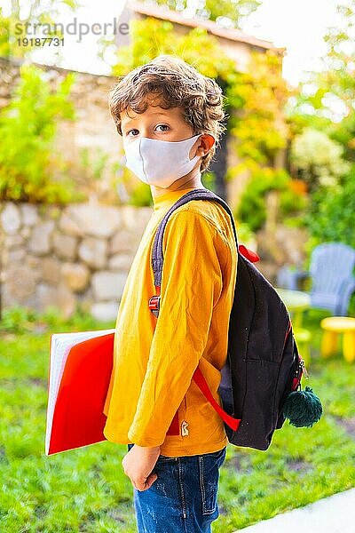 Kaukasisches Kind mit Gesichtsmaske  bereit für die Rückkehr zur Schule. Neue Normalität  soziale Distanz  Coronaviruspandemie Covid 19. Orangefarbenes TShirt Rucksack und ein Notizblock in der Hand