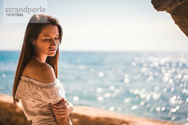 Lebensstil eines jungen rothaarigen  weiß gekleideten Mädchens neben einem Felsen am Meer