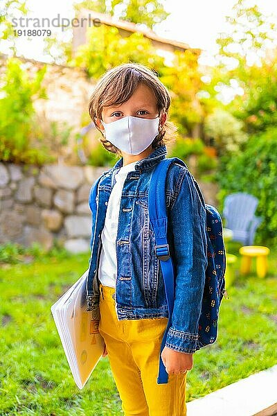 Kaukasisches Kind mit Gesichtsmaske  bereit für die Rückkehr zur Schule. Neue Normalität  soziale Distanz  Coronaviruspandemie Covid 19. Mit Jeansjacke  Rucksack und Notizblock in der Hand