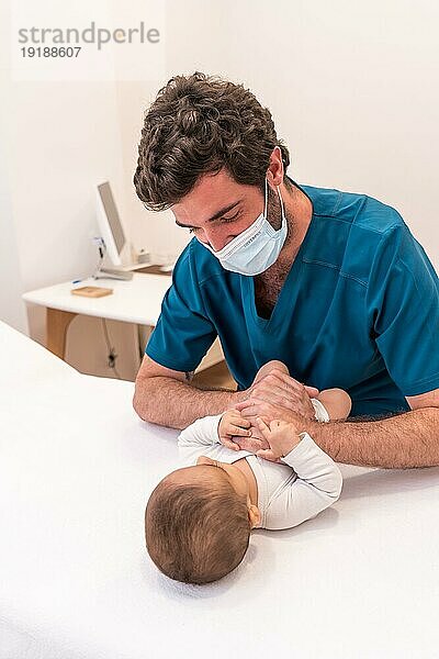 Frontalaufnahme eines jungen Arztes mit Gesichtsmaske  der ein süßes Neugeborenes in einer Klinik untersucht