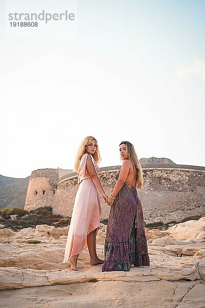 Ein paar kaukasische Mädchen mit rosa Kleidern  im Sommer am Playazo de Rodalquilar in Cabo de Gata neben der Burg im Sommer