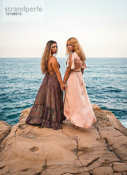 Ein paar kaukasische Mädchen in schönen Kleidern  die Hände in einer zärtlichen Art und Weise durch das Meer im Sommer am Strand