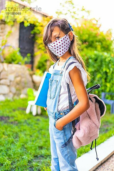 Kaukasisches Mädchen mit Gesichtsmaske  bereit  wieder zur Schule zu gehen. Neue Normalität  soziale Distanz  Coronaviruspandemie Covid 19. Jacke  Rucksack und ein blauer Block für Notizen in der Hand