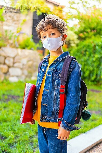 Kaukasisches Kind mit Gesichtsmaske  bereit für die Rückkehr zur Schule. Neue Normalität  soziale Distanz  Coronaviruspandemie Covid 19. Jacke  Rucksack und ein roter Block für Notizen in der Hand