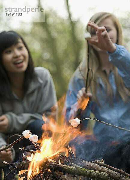 Zwei lächelnde Mädchen im Teenageralter rösten Marshmallows über einem Lagerfeuer
