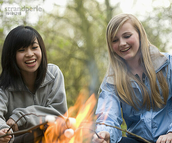 Zwei lächelnde Mädchen im Teenageralter rösten Marshmallows über einem Lagerfeuer