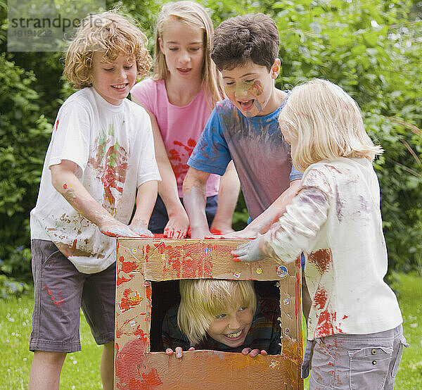 Mit Aquarellfarbe bedeckte Kinder spielen in einem Garten  eines davon liegt in einem Karton