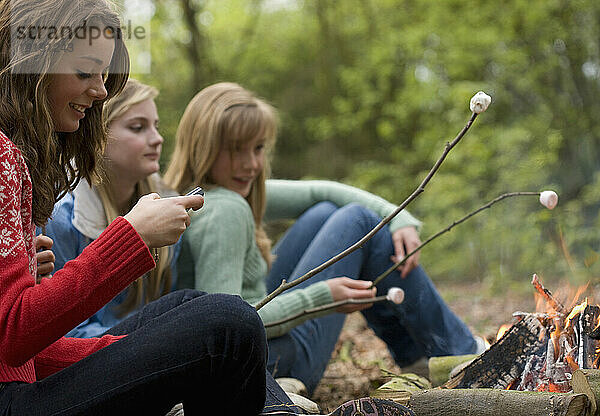 Teenager-Mädchen rösten Marshmallows über dem Lagerfeuer  eines benutzt ein Mobiltelefon