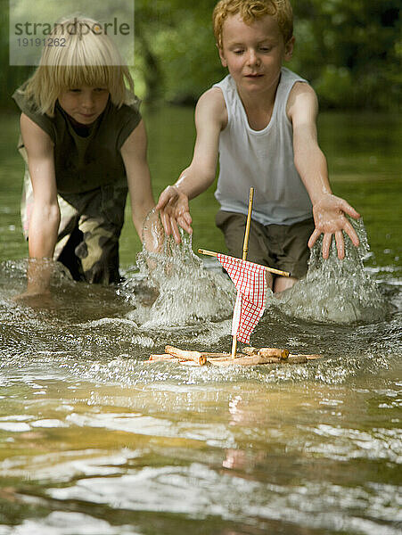 Zwei kleine Jungen in einem Fluss spritzen Wasser auf ein Modellfloß zu
