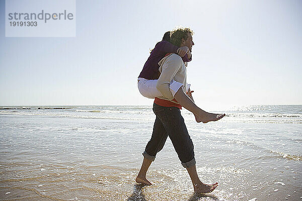 Mann trägt Frau auf dem Rücken und geht am Strand spazieren