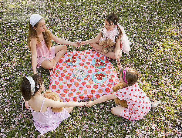 Vier junge Mädchen sitzen um eine bunte Decke herum und halten Händchen und feiern eine Party
