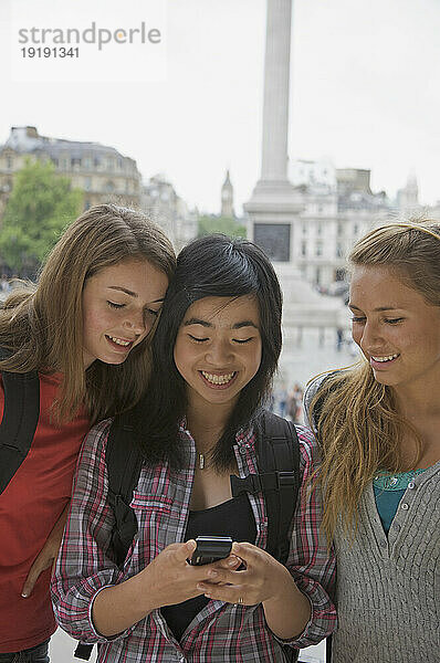 Drei lächelnde Mädchen im Teenageralter  die auf dem Londoner Trafalgar Square ein Mobiltelefon benutzen