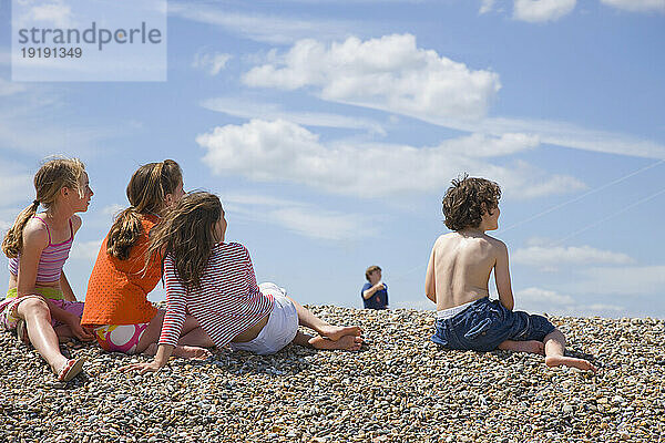 Kinder sitzen am Strand und beobachten einen kleinen Jungen  der einen Drachen steigen lässt