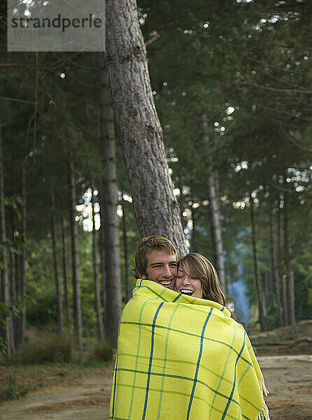 Junges Paar steht lachend in einem Wald  in eine Decke gehüllt