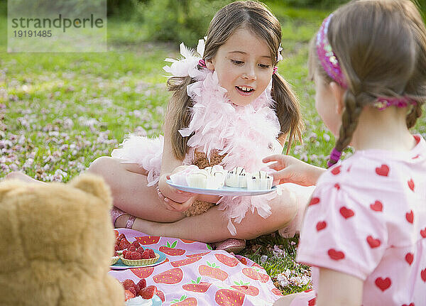 Junges Mädchen sitzt in einer Gartenwiese und trägt eine rosa Federboa und bietet einer Freundin Cupcakes an