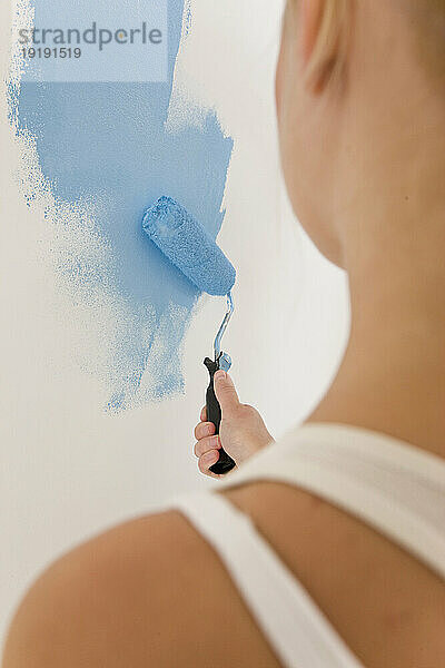 Rückansicht einer Frau  die einen Pinsel hält und eine weiße Wand mit blauer Farbe bemalt