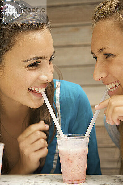 Zwei lächelnde Mädchen im Teenageralter trinken Milchshake mit einem Strohhalm aus demselben Glas