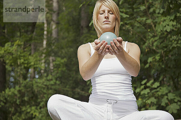 Junge Frau in Meditation sitzt im Schneidersitz an einem Wald und hält eine Kugel in ihren Händen